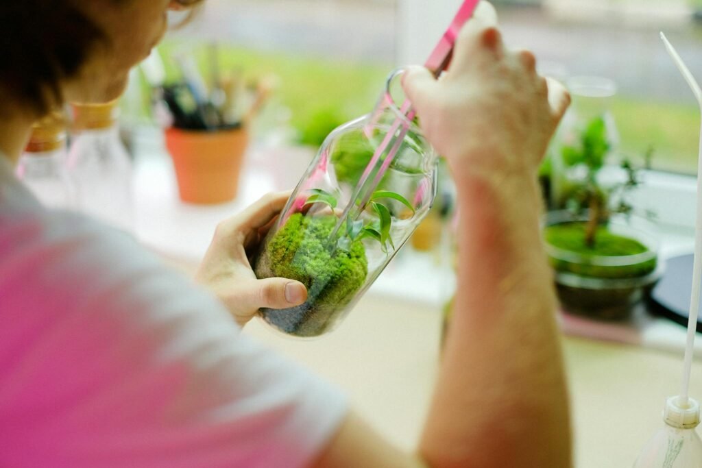 Man using tongs to arrange a moss terrarium in a glass jar under natural light.